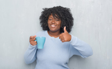 Sticker - Young african american woman over grey grunge wall drinking a cup of coffee happy with big smile doing ok sign, thumb up with fingers, excellent sign