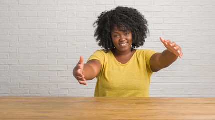 Canvas Print - Young african american woman sitting on the table at home looking at the camera smiling with open arms for hug. Cheerful expression embracing happiness.