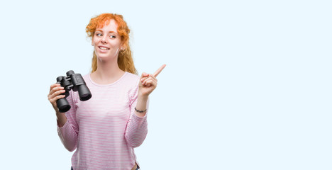 Poster - Young redhead woman looking through binoculars very happy pointing with hand and finger to the side