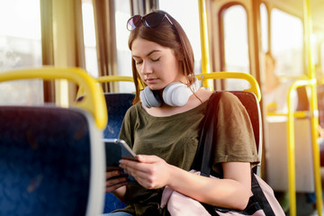 Wall Mural - Serious young student girl sitting in a bus and looking at her telephone with serious look. Holding headphones around her neck.