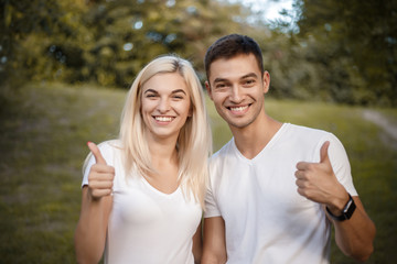 Wall Mural - Happy young cute loving couple standing on grass in nature green park with beautiful sundown light. Looking camera showing thumbs up gesture.