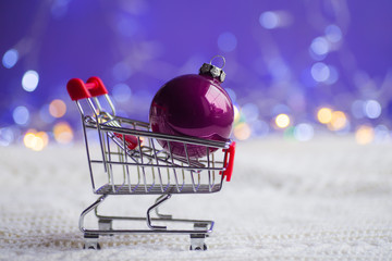 Crimson Christmas ball in small supermarket trolley on white knitted fabric with garland lights on purple bokeh background.