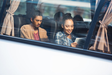 Wall Mural - smiling asian woman using digital tablet while her mixed race boyfriend sitting behind during trip on travel bus