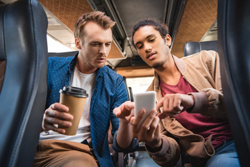 Wall Mural - young mixed race man showing smartphone to his male friend with paper coffee cup during trip on bus