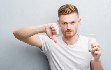 Wall Mural - Young redhead man over grey grunge wall drinking a glass of milk with angry face, negative sign showing dislike with thumbs down, rejection concept