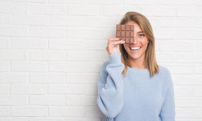 Beautiful young woman over white brick wall eating chocolate bar with a happy face standing and smiling with a confident smile showing teeth