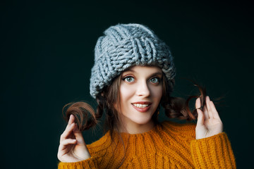 Close up studio portrait of young beautiful happy smiling girl wearing stylish gray beanie hat and yellow sweater, holding her hair, braids posing on dark background. Copy, empty space for text