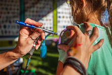 Young Girl Getting An Airbrush Stencil Temporary Tattoo In A Family Festival Outdoors - Closeup Picture With Many Different Tattoo Designs Displayed In The Blurry Background