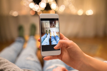 leisure, hygge and christmas concept - close up of couple taking foot selfie by smartphone and garland lights at home