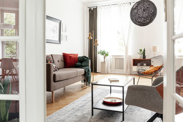 Table on carpet between couch and armchair in bright loft interior with lamp and plants. Real photo