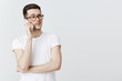© Cookie Studio - Waist-up shot of bossy and busy young creative male entrepreneur making appointment via phone holding smartphone near ear gazing uncertain and doubtful right wearing glasses over grey wall