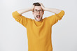 © Cookie Studio - Man yelling cannot control panic and stress holding hands on head open mouth and screaming out loud from distress and anxiety standing troubled and upset in trendy yellow sweater over white background