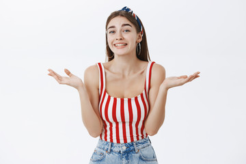 Sorry no care at all. Portrait of clueless chill silly european female in striped top and headband shrugging with palms spread aside smiling with sorry look being unsure and unaware what happening