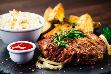 Grilled steak with baked potatoes and vegetables served on black stone on wooden table