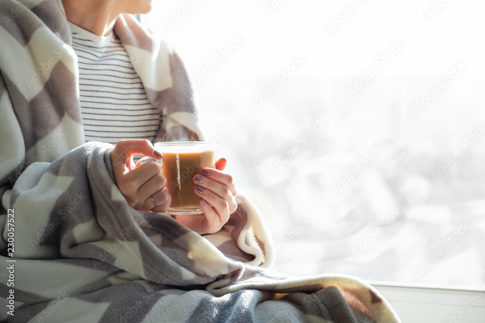 Hot steaming drink in woman's hands. Female covered in throw blanket sits by the window with cup of hot cocoa - obrazy, fototapety, plakaty 