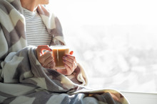 Hot Steaming Drink In Woman's Hands. Female Covered In Throw Blanket Sits By The Window With Cup Of Hot Cocoa