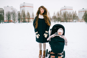 Wall Mural - Fashionable stylish young mother rest with her daughter sitting in stroller in park outdoor at winter. Motherhood happiness. Children - flowers of life.  Beautiful girl with her baby winter portrait.