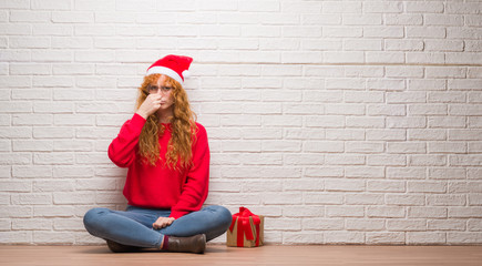 Wall Mural - Young redhead woman sitting over brick wall wearing christmas hat smelling something stinky and disgusting, intolerable smell, holding breath with fingers on nose. Bad smells concept.