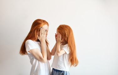 Ginger haired Mom and little daughter, so much alike, both with long loose red hair down, looking at each other with great surprise or amazement, touching with hands their cheeks in disbelief