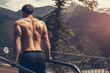 © alfa27 - Backside view of muscular fair-haired european man in black shorts and naked top doing dips on parallel bars . Outdoor workout with blue sky and green mountains on the background and panoramic views.