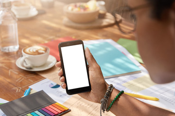 Back view of black woman holds modern cellular with mock up screen to paste design content, poses in cafeteria, studies papers, drinks coffee, uses mobile phone application. Empty copy space