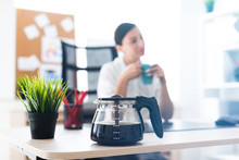 Photo With Depth Of Field, Highlighted Focus On The Coffee Pot. A Young Girl Sitting At A Table In The Office And Holding A Cup.