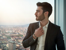 Portrait Of Stylish Young Man Wearing Business Suit, Standing Next To Window Overlooking Modern European City