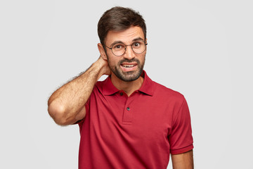 Wall Mural - Photo of puzzled man with stubble, scratches head, looks in bewilderment, wears round spectacles for good vision, dressed in casual red t shirt, models against white background has confused expression