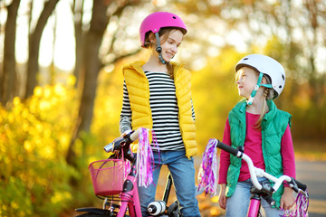 Cute sisters riding bikes in a city park on sunny autumn day. Active family leisure with kids. Children wearing safety hemet while riding a bicycle.