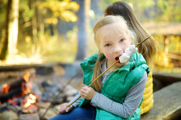 Wall Mural - Cute little girl roasting marshmallows on stick at bonfire. Child having fun at camp fire. Camping with children in fall forest.