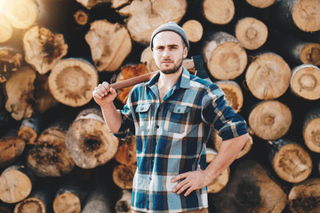 Wall Mural - Strong bearded lumberman wearing plaid shirt holds axe on his shoulder on background of sawmill