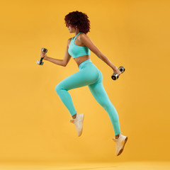 Wall Mural - Strong athletic, woman sprinter or runner, running on yellow background with dumbbells wearing sportswear. Fitness and sport motivation.