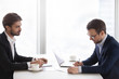 © fizkes - Two caucasian men in suits sitting opposite each other and discussing or evaluating or planning project. Partners or customer and seller are calculate or analyze contract information before signing