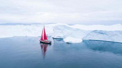 Wall Mural - beautiful red sailboat in the arctic next to a massive iceberg showing the scale - aerial view as vi