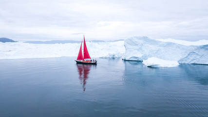 Wall Mural - beautiful red sailboat in the arctic next to a massive iceberg showing the scale - aerial view as vi