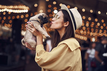 Kissing Puppy. Happy Young Pretty Lady Standing Outdoors With Hat On Her Head And Kissing Cute Puppy In Her Hands