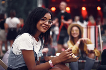 Wall Mural - Portrait of beautiful young lady in glasses holding food and looking at camera with smile. Stage and people on blurred background