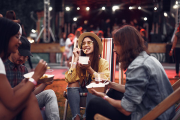 Wall Mural - Portrait of cheerful young lady in hat sitting on folding chair and eating french fries while handsome bearded guys holding beer. Stage and crowd on blurred background