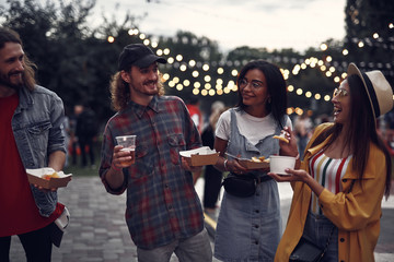 Wall Mural - Waist up portrait of stylish young people enjoying meal while standing on the street and chatting. They looking at each other and smiling