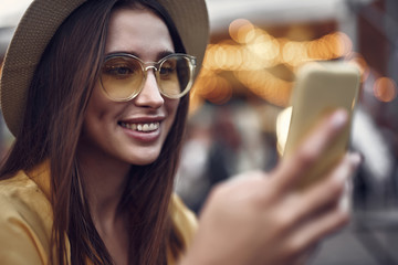 Wall Mural - Surfing internet. Close up portrait of hipster young lady in hat holding smartphone and looking at display with smile. Focus on woman face