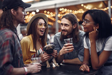 Waist Up Portrait Of Stylish Hipster Friends Enjoying Drinks While Standing At The Table. Cheerful Girl In Hat Holding Little Cute Dog