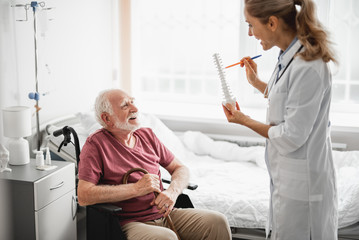 Side view portrait of smiling bearded gentleman looking at young lady in white lab coat while she holding anatomical model of spine and pointing at it. Patient holding walking stick