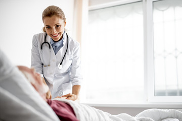 Waist up portrait of beautiful young woman in white lab coat holding hand of old man and looking at him with smile. Copy space on right side