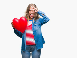 Wall Mural - Beautiful young blonde woman holding heart valentine over isolated background with happy face smiling doing ok sign with hand on eye looking through fingers