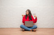 © Krakenimages.com - Young brunette woman sitting on the floor over white brick wall paying holding dollars pointing and showing with thumb up to the side with happy face smiling