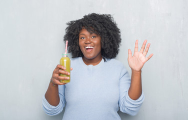 Poster - Young african american woman over grey grunge wall drinking detox green juice very happy and excited, winner expression celebrating victory screaming with big smile and raised hands