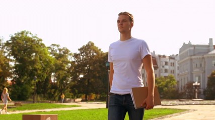 Leinwandbilder - caucasian student walking on the street. young man with blond hair holding notebook going to class slow motion