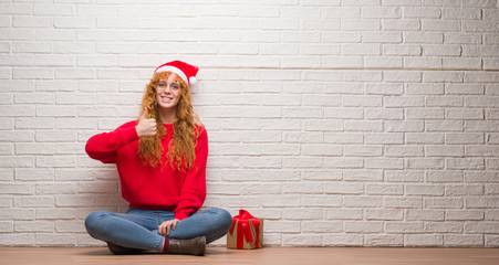 Poster - Young redhead woman sitting over brick wall wearing christmas hat doing happy thumbs up gesture with hand. Approving expression looking at the camera with showing success.