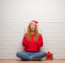 Poster - Young redhead woman sitting over brick wall wearing christmas hat amazed and surprised looking up and pointing with fingers and raised arms.