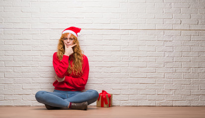 Wall Mural - Young redhead woman sitting over brick wall wearing christmas hat looking confident at the camera with smile with crossed arms and hand raised on chin. Thinking positive.